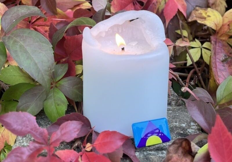 A lit candle among autumn leaves with a meningitis flag badge resting against it.